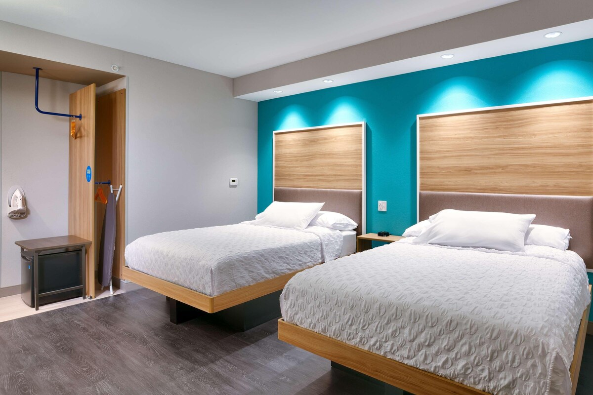 A hotel room with two beds, a wooden headboard, a small bedside table, and a wall-mounted heater.