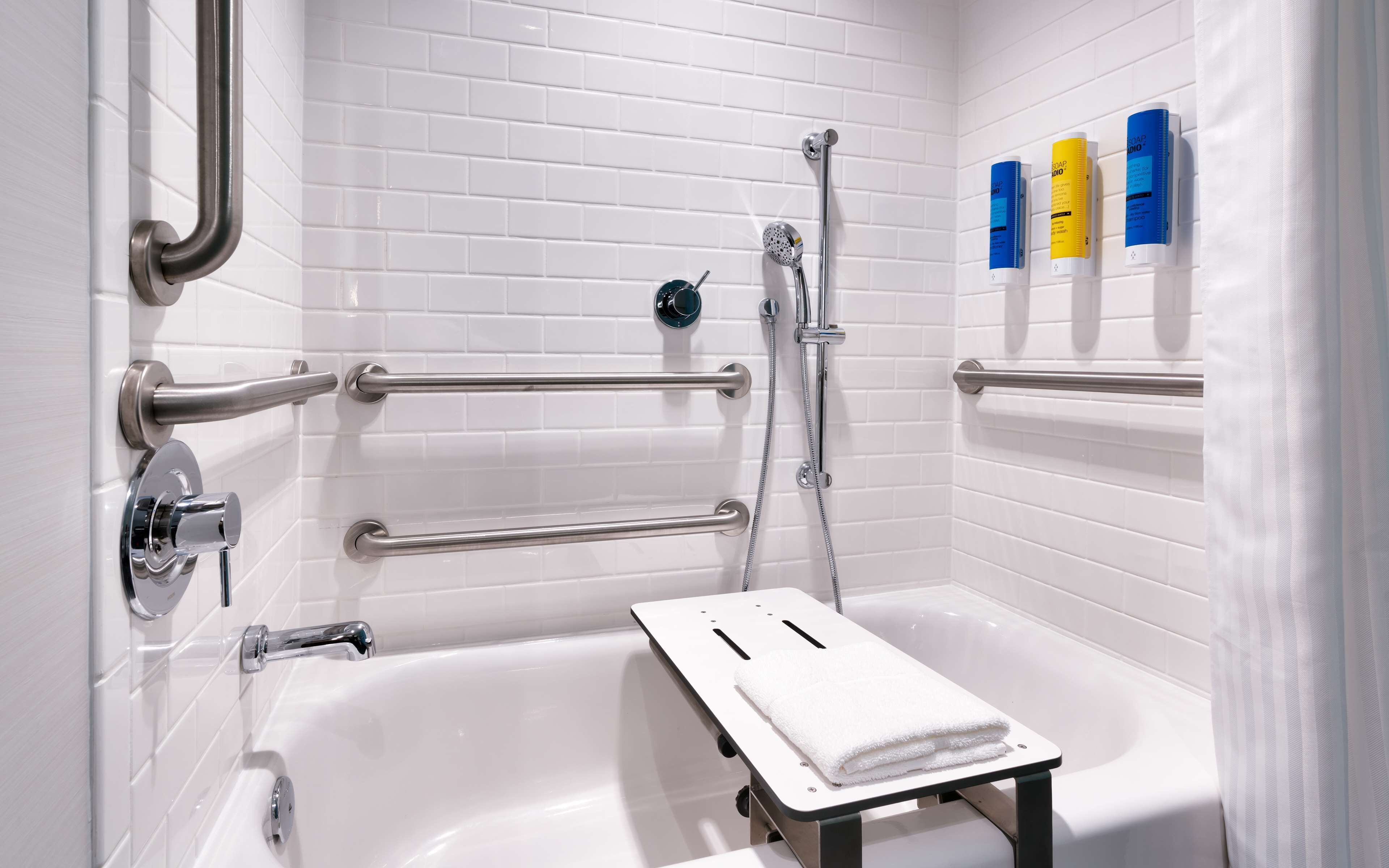 A modern bathroom with white tiled walls, a white bathtub, and a shower area. There are grab bars installed for safety, and a towel rack with soap dispensers is visible.