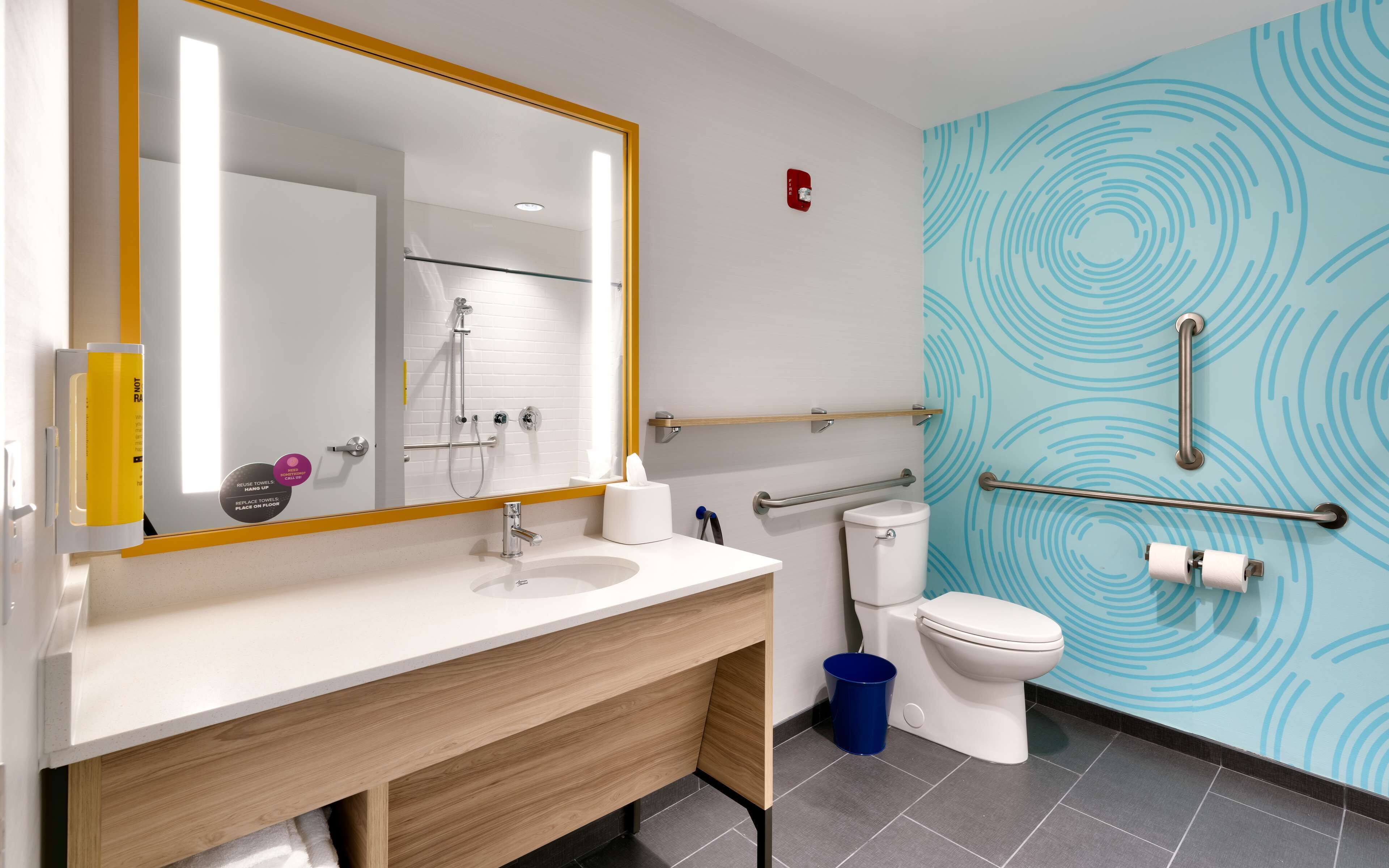 A modern bathroom with a white sink, mirror, and toilet. There are handrails and a blue trash bin.