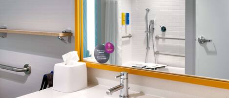 A bathroom with a white sink, a mirror, and a shower. There are handrails and a towel dispenser.