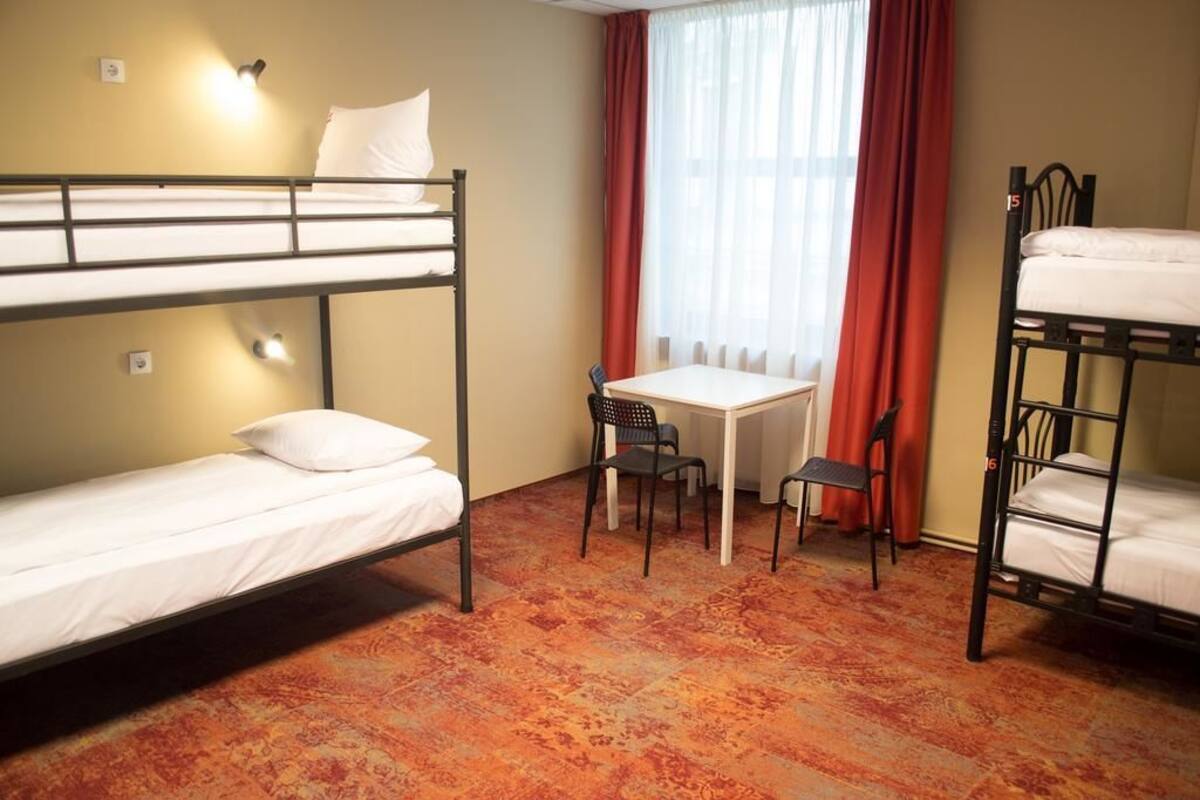 A dormitory room with bunk beds, a small table, two chairs, and a window with curtains.