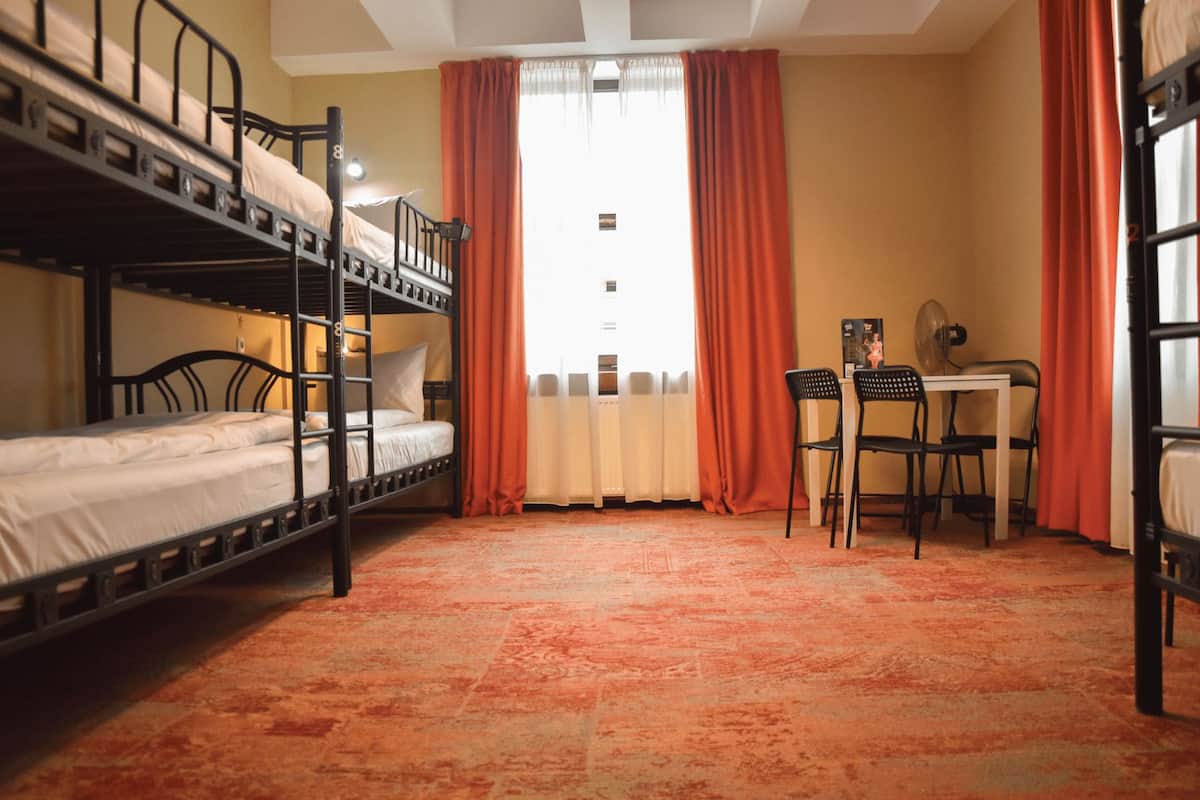 A dormitory room with bunk beds, a desk, two chairs, and a window with curtains.