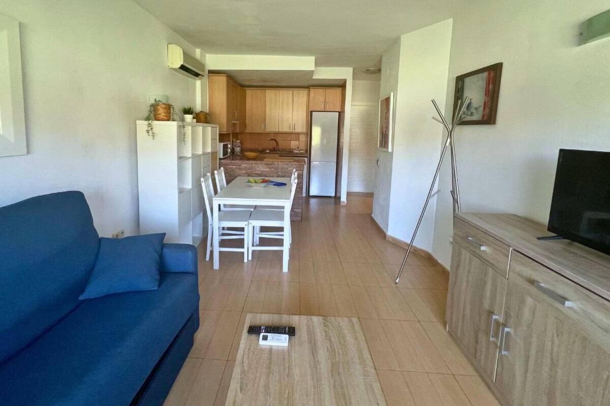 Una sala de estar moderna con un sofá azul, un comedor con una mesa y sillas blancas, una cocina con armarios de madera y un televisor sobre un mueble de madera.