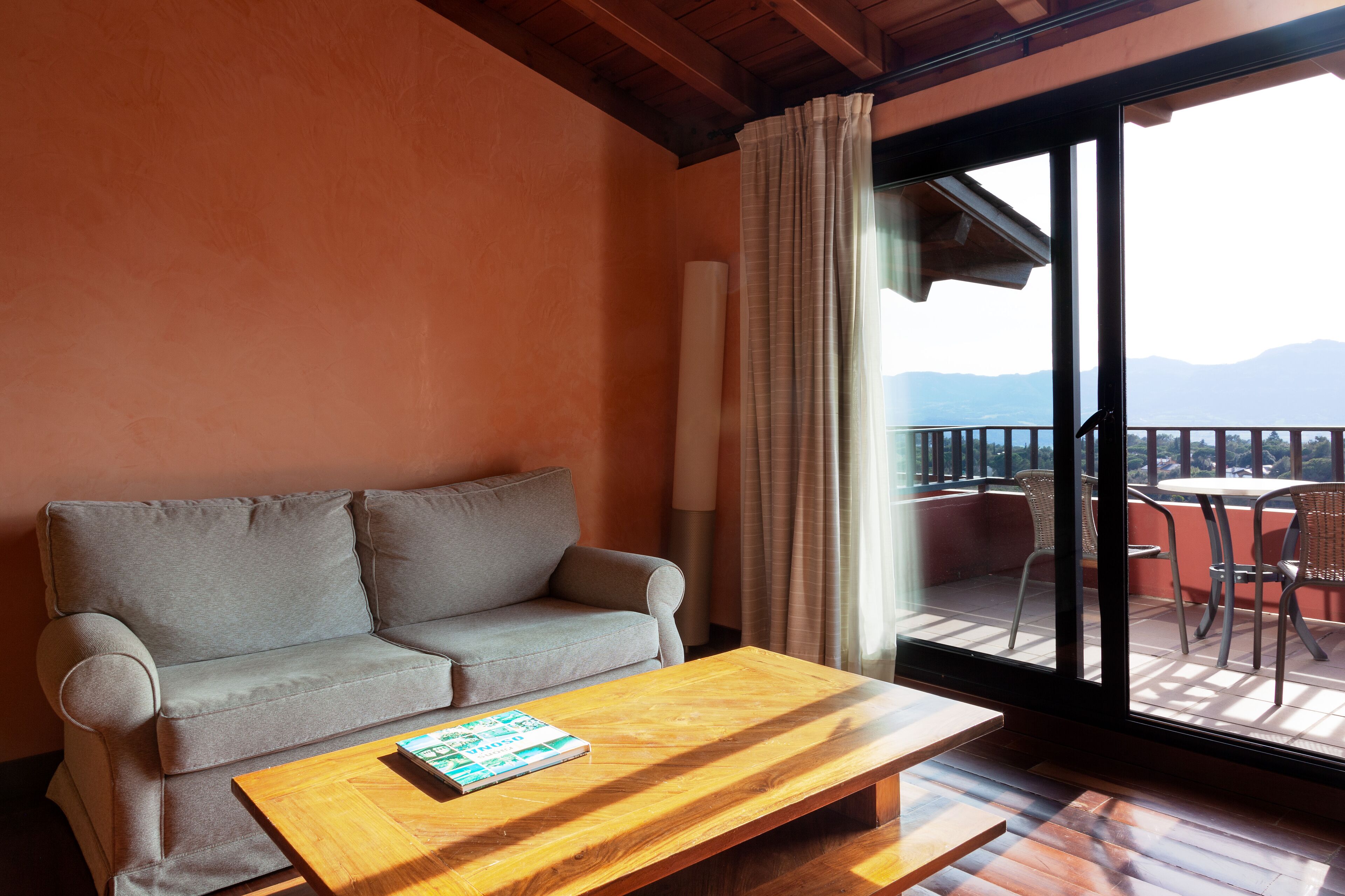 A room with a sofa, a wooden coffee table, and a view of a balcony with chairs.
