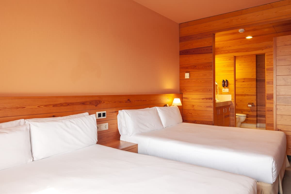 Habitación de hotel con dos camas, paneles de madera, una mesita de noche con lámpara y un espejo que refleja el baño.
