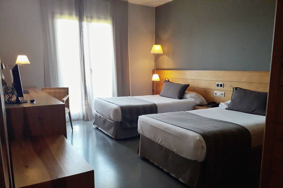 Habitación de hotel con dos camas, un escritorio y una silla. Tiene dos lámparas de noche y una ventana con cortinas.