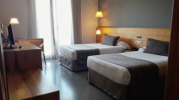 A hotel room with two beds, a desk, and a chair. There are two bedside lamps and a window with curtains.