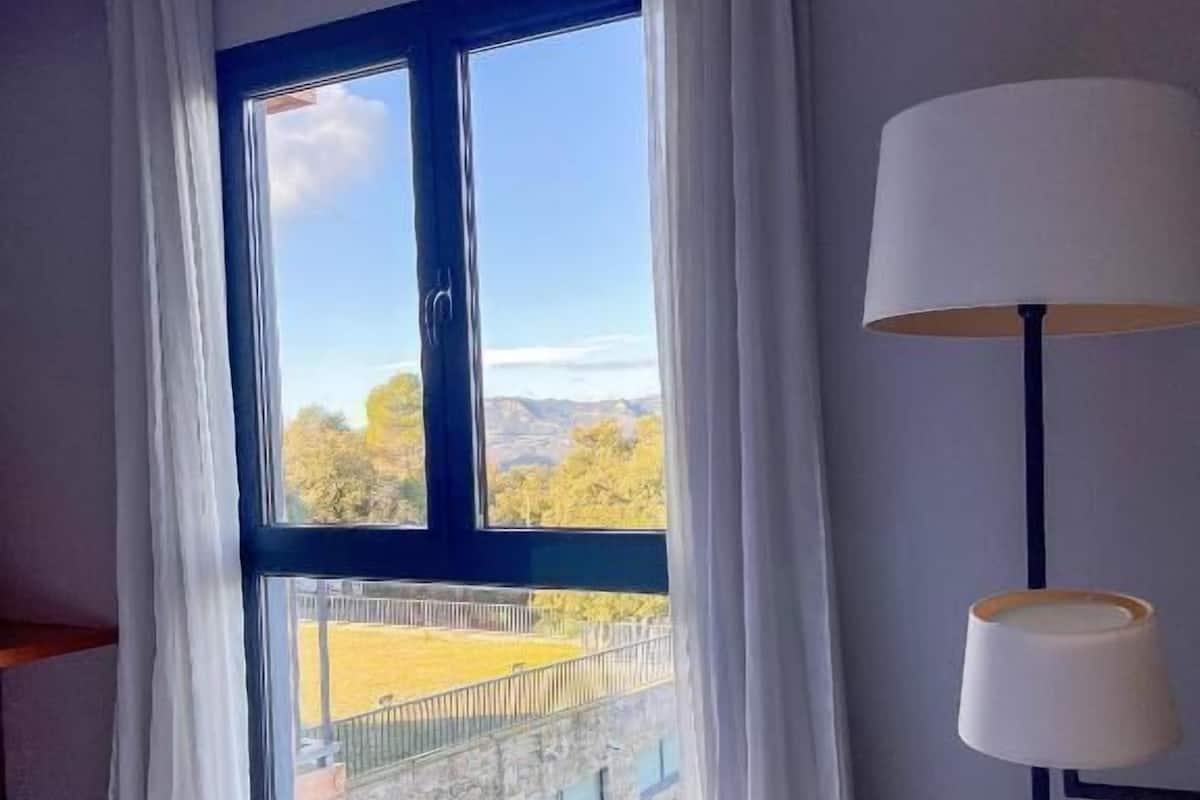 Habitación de hotel con un ventanal grande que ofrece vistas a un edificio y árboles. Hay una cama con ropa blanca y una lámpara sobre una mesita de noche.