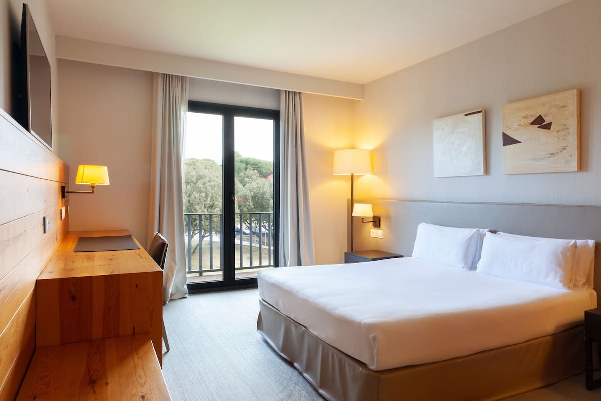 Habitación de hotel con cama, escritorio, silla, televisión, balcón y dos lámparas fijadas a la pared.