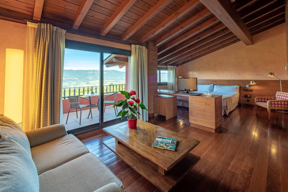 Una habitación de hotel moderna con techo de madera, balcón con vistas, zona de comedor y un rincón de descanso acogedor.
