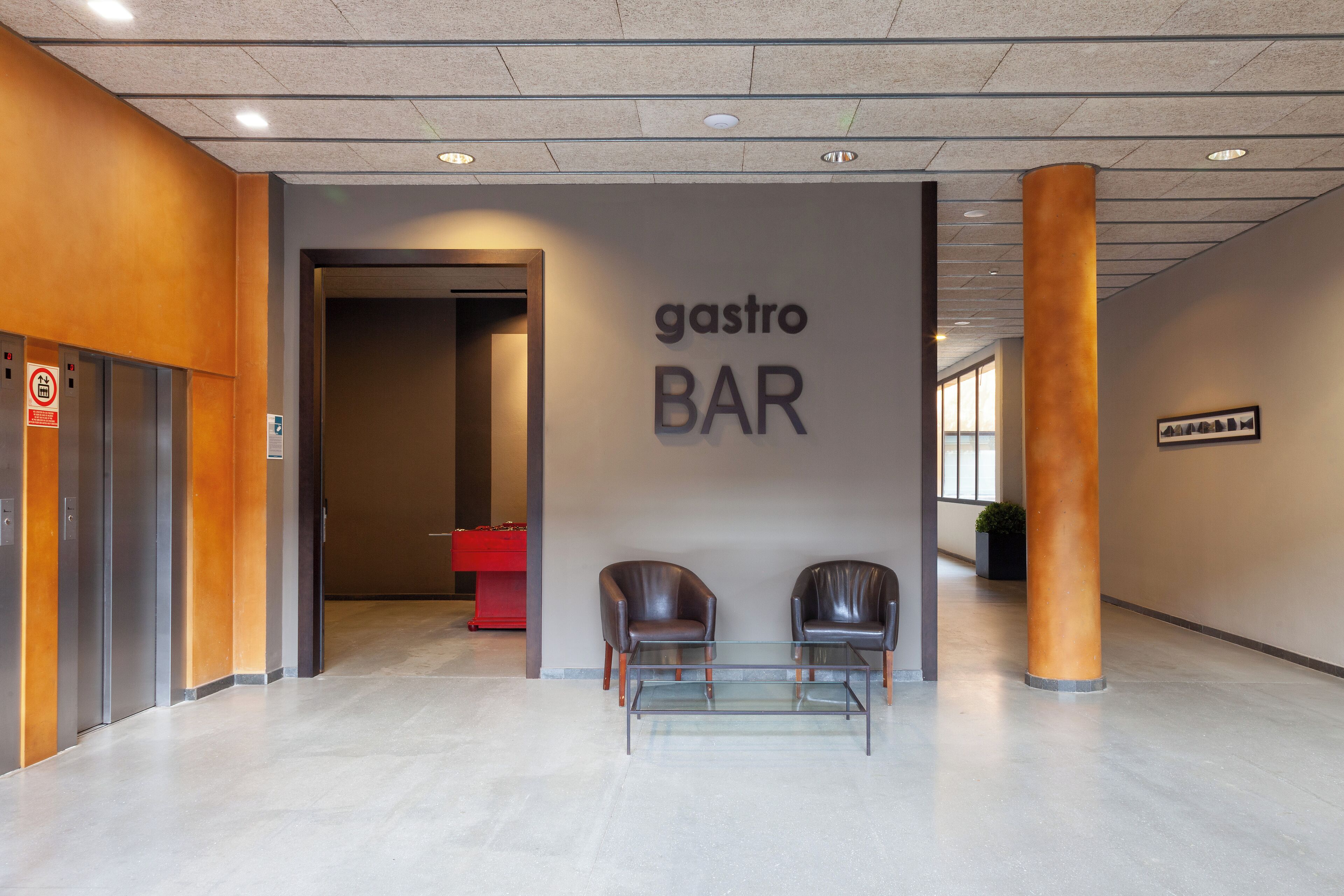 Un interior modern amb un cartell de 'gastro BAR', dues butaques de pell, una taula de vidre i un taulell vermell al fons.