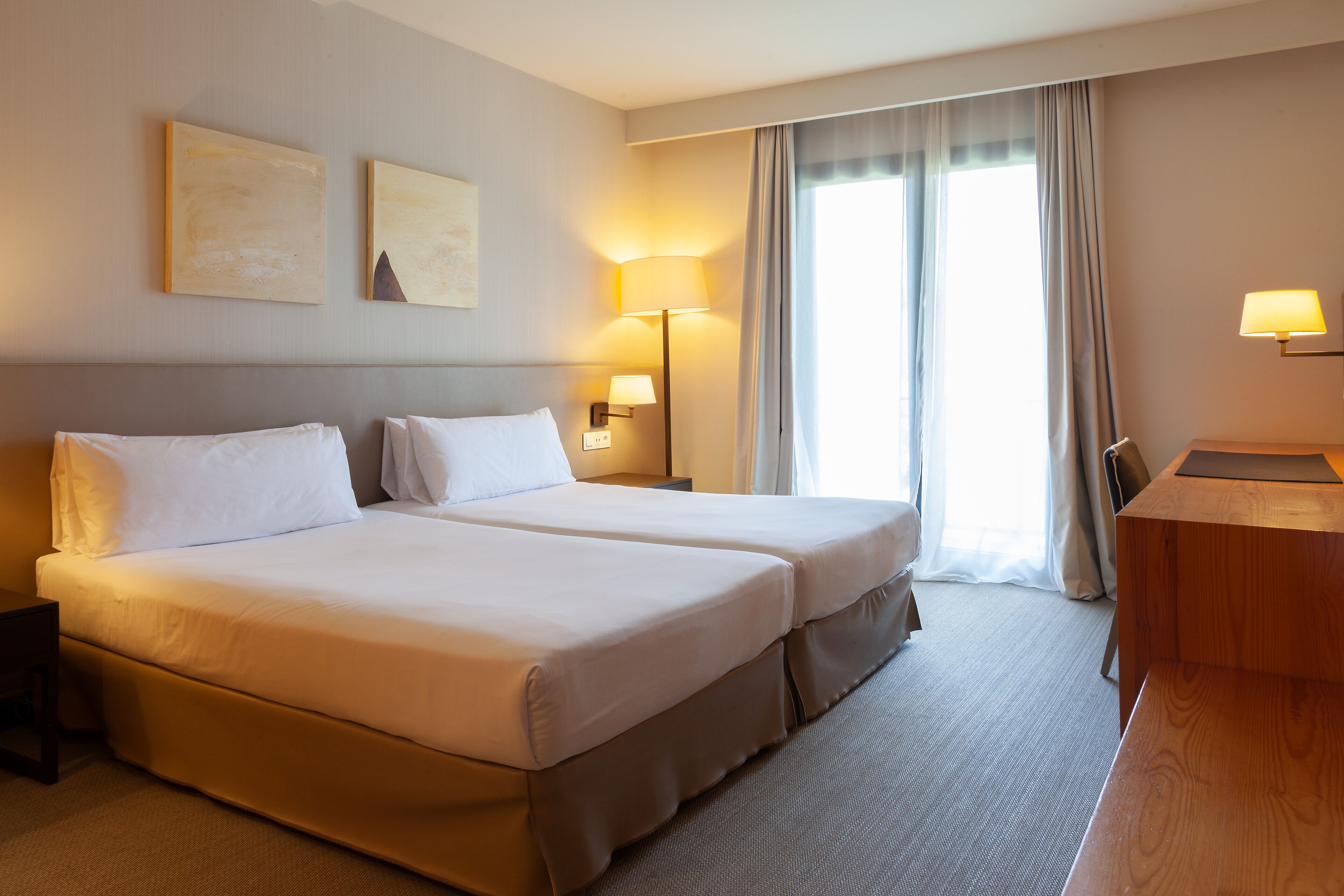 A hotel room with two beds, a desk, and a lamp.
