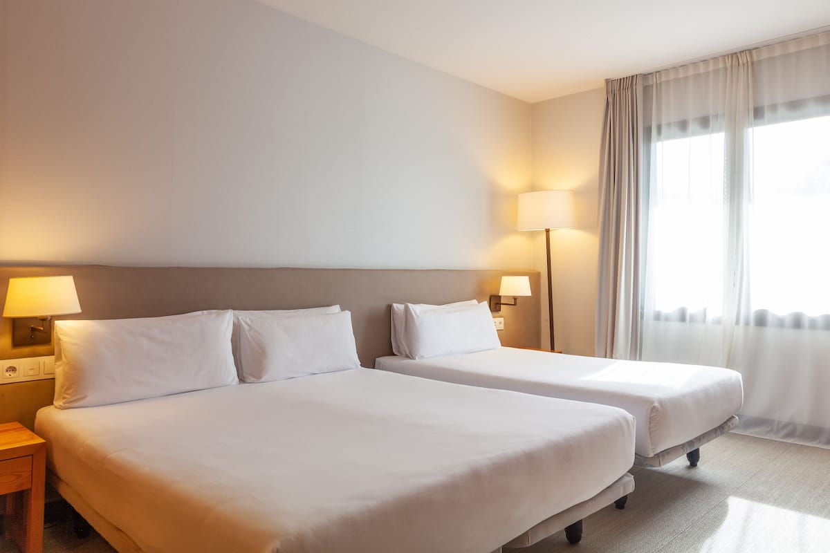 Habitación de hotel con dos camas individuales, una mesita de noche con lámpara y una ventana con cortinas translúcidas.