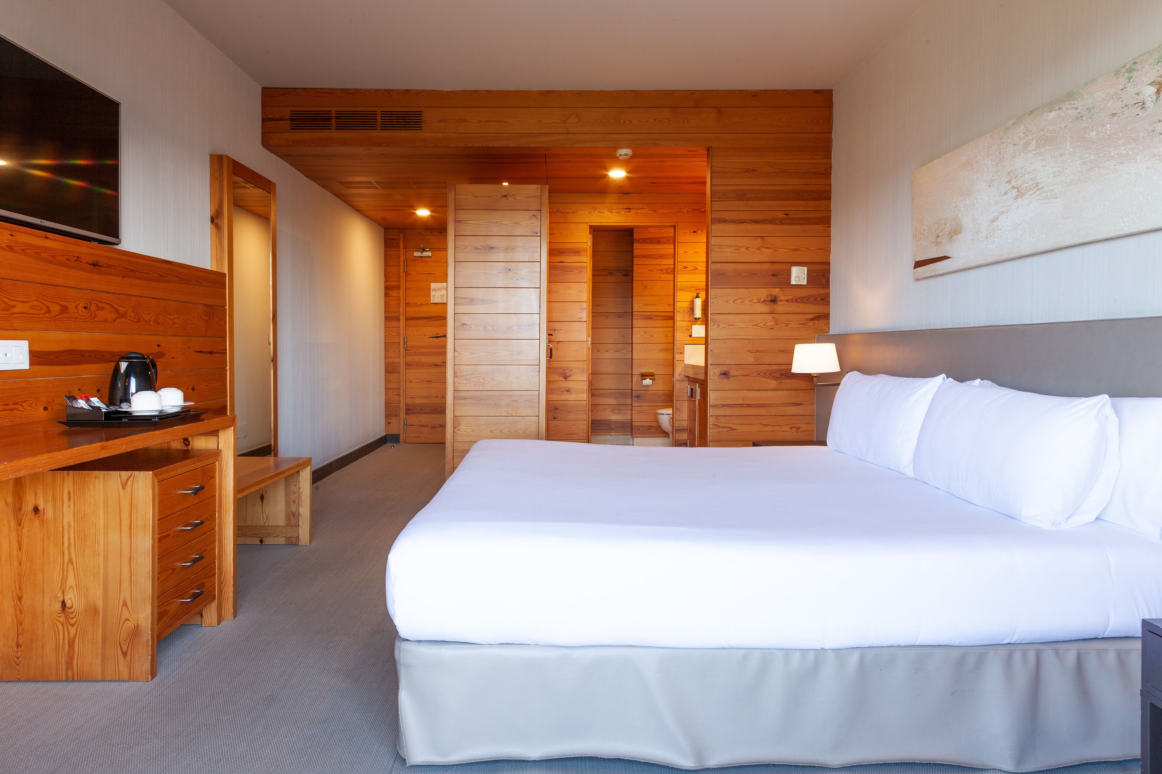 A hotel room with a large bed, wooden paneling, a desk with a coffee maker, and a TV on the wall.