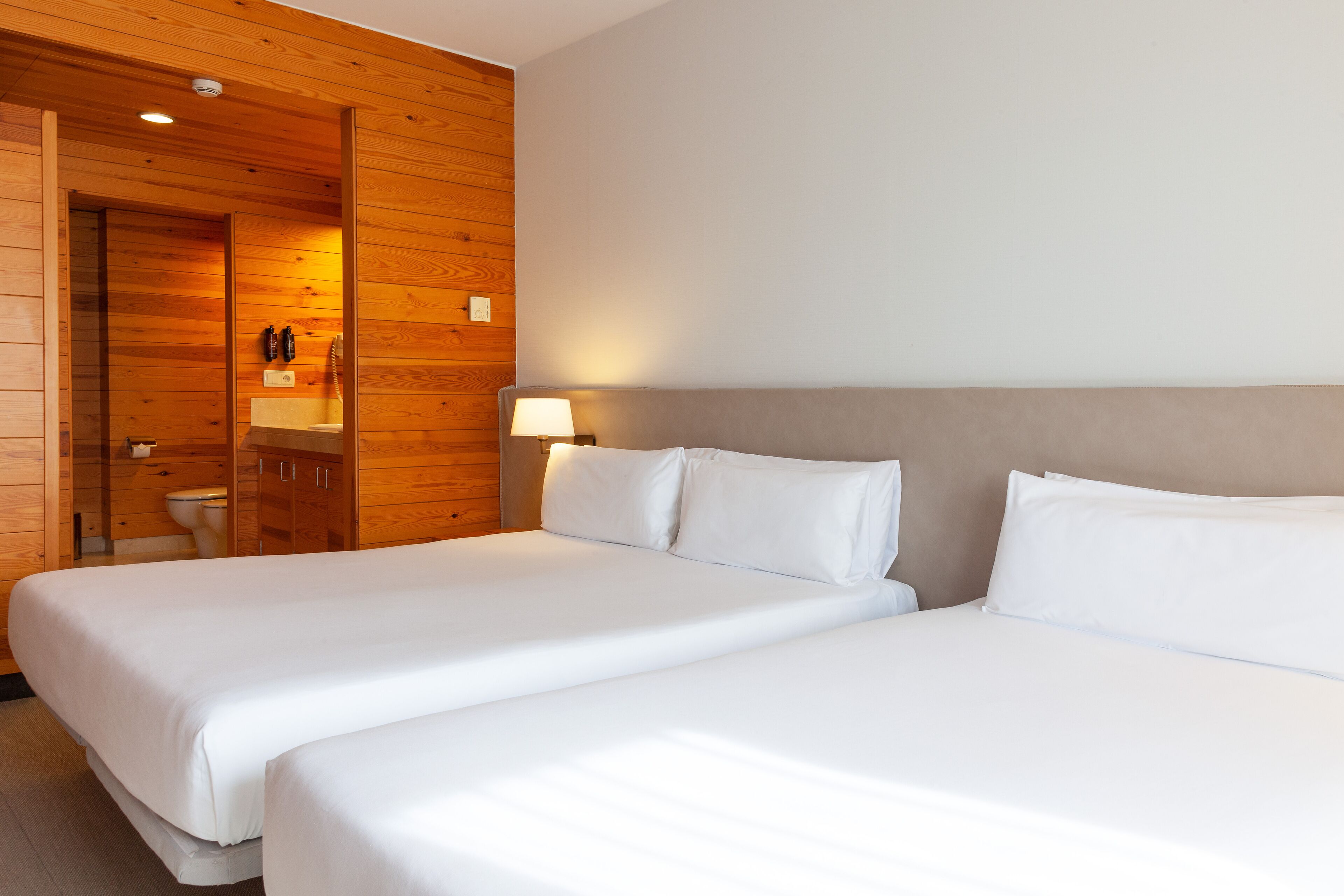 Habitación de hotel con dos camas, paneles de madera, un baño con inodoro y lavabo, y una mesita de noche con lámpara.