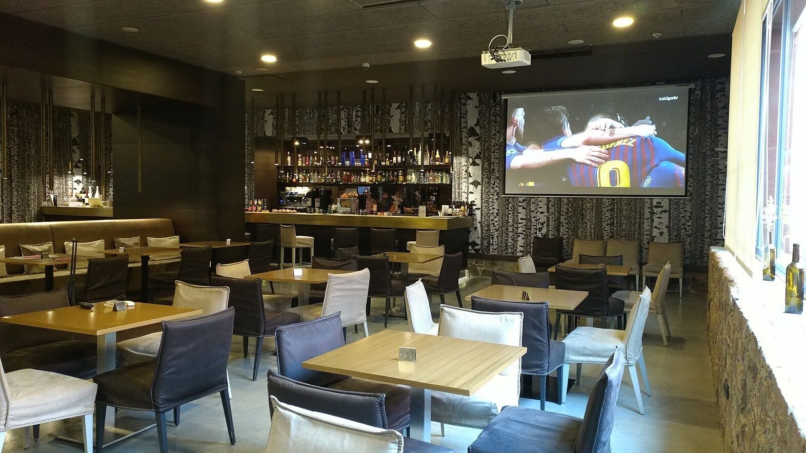 A restaurant with multiple tables and chairs, a bar area with shelves of bottles, and a large screen displaying a sports event.