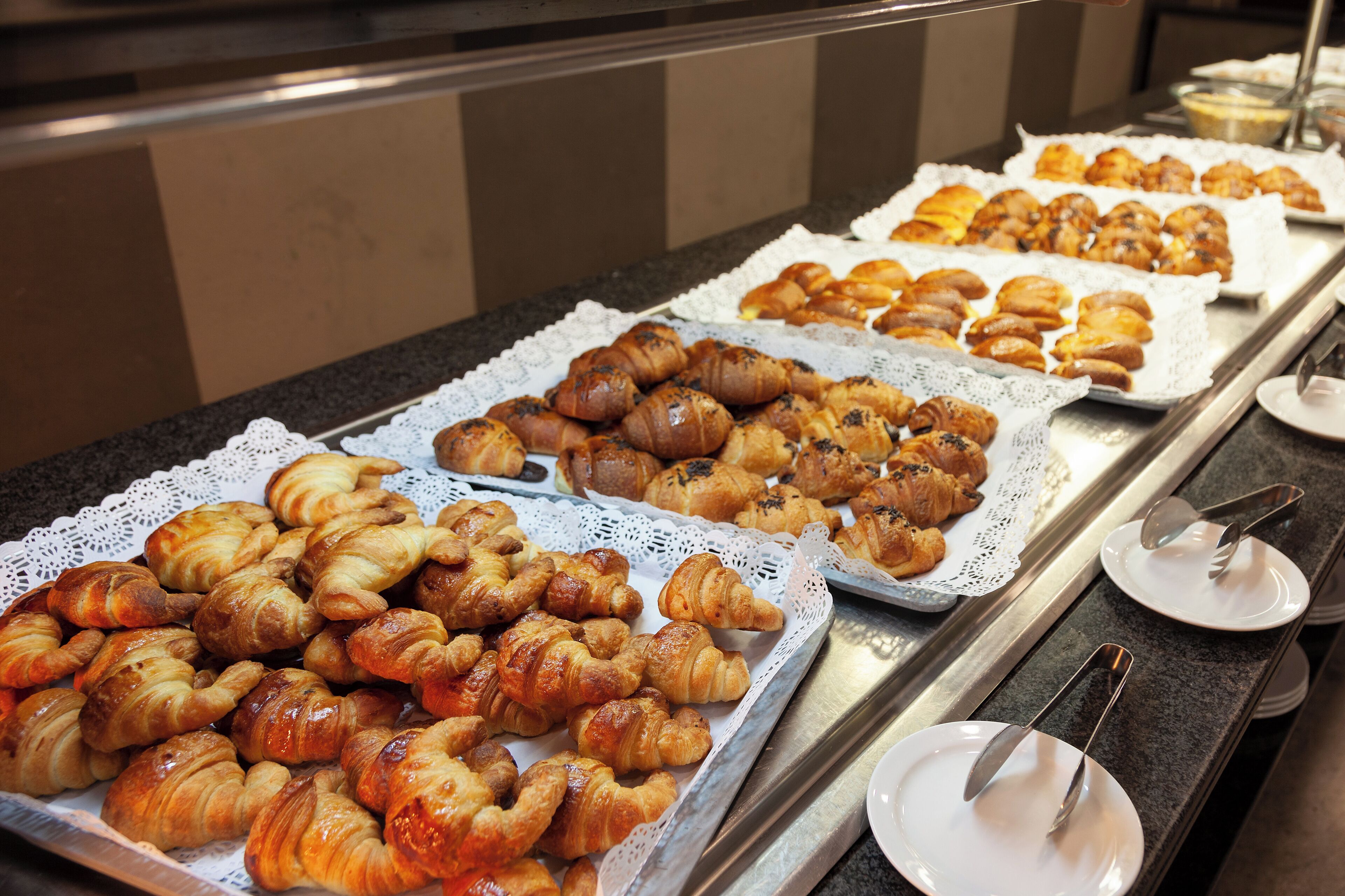 Un bufé con diversas pastas y panes, incluyendo cruasanes y muffins, dispuestos en bandejas.
