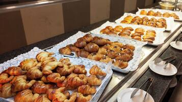 Un bufé con diversas pastas y panes, incluyendo cruasanes y muffins, dispuestos en bandejas.