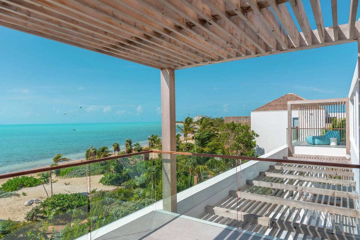 A balcony with a view of the ocean and beach, wooden railings, and a staircase leading to a higher floor.