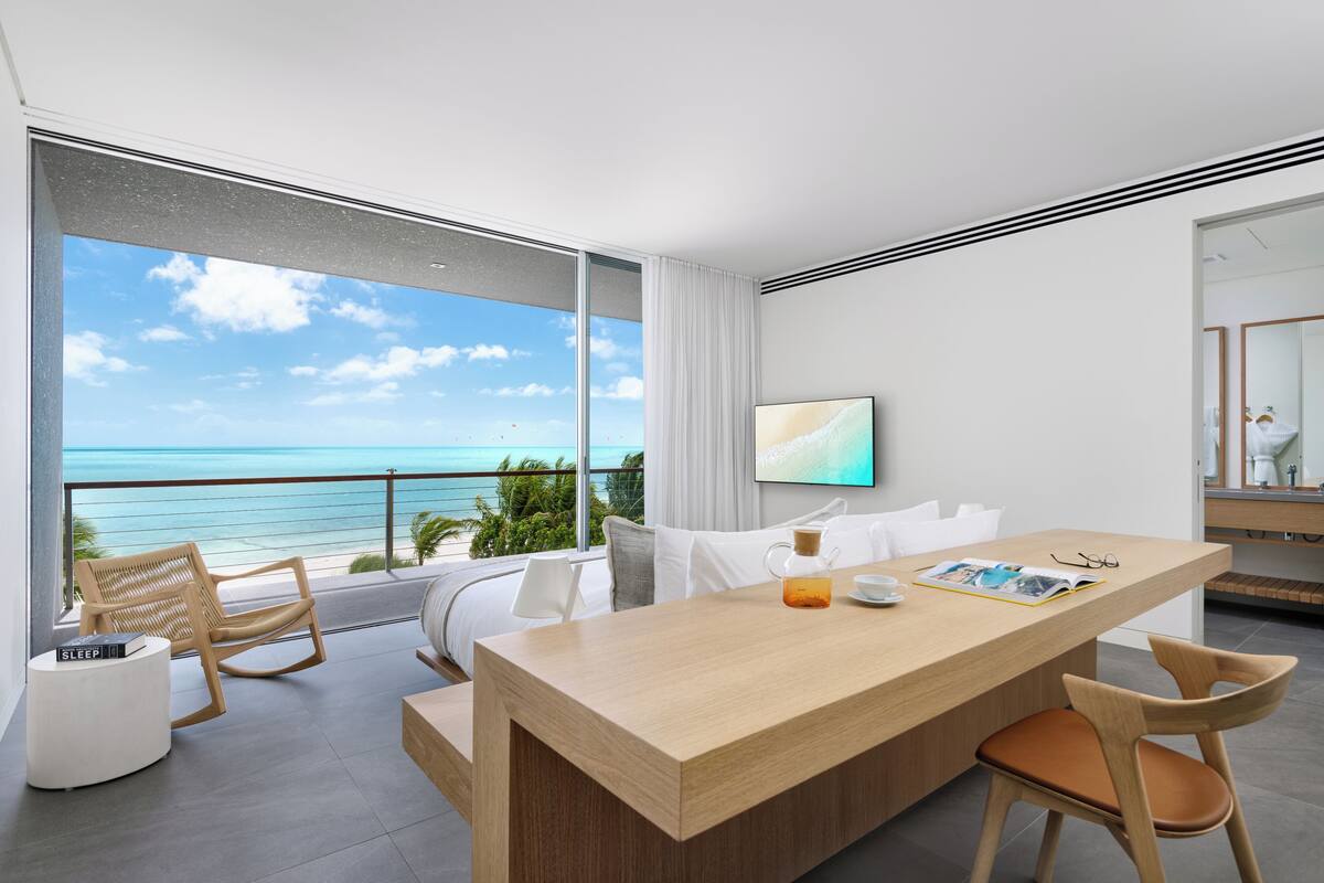 A modern hotel room with a large balcony, a wooden dining table, and a view of the ocean.