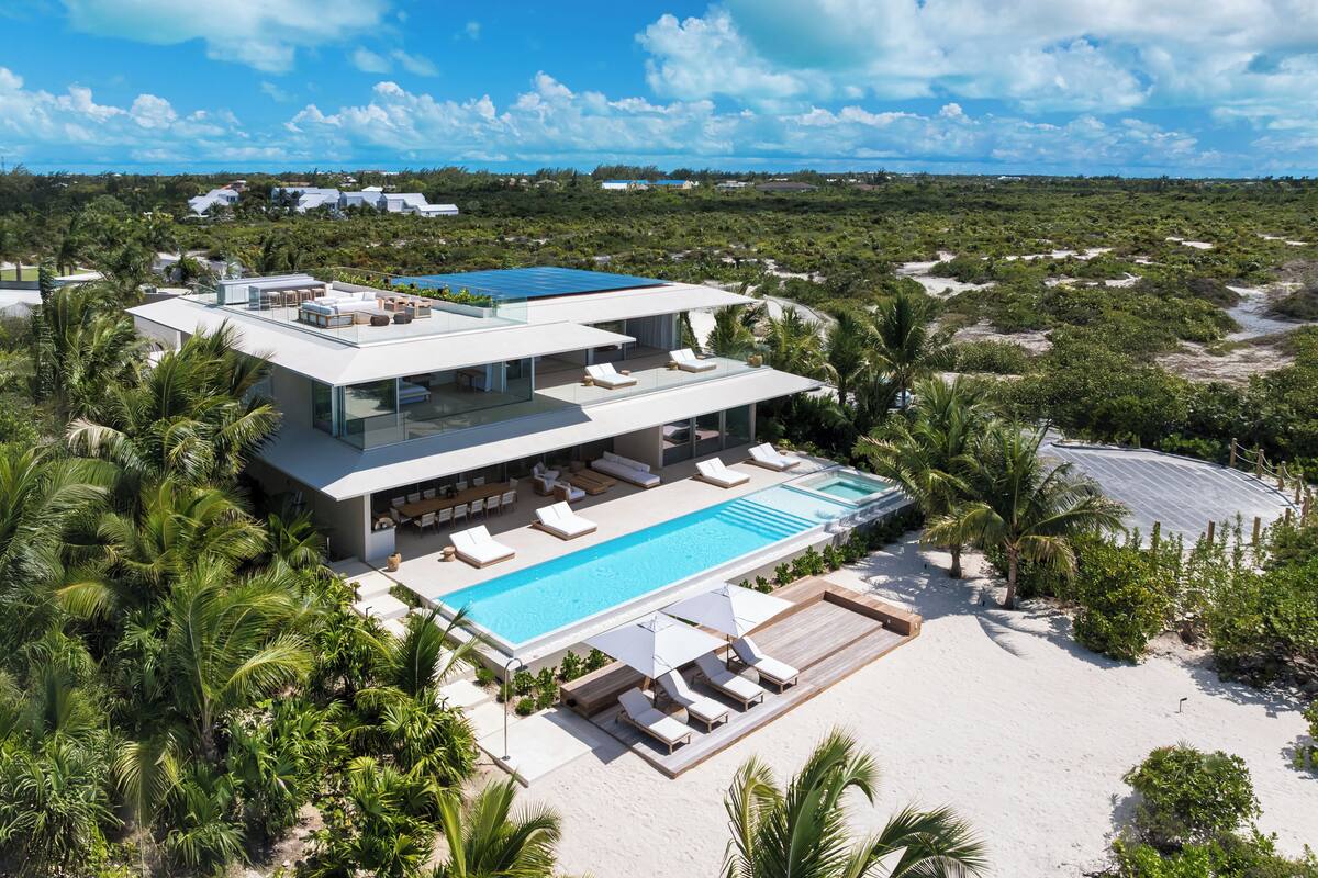 A modern multi-story house with a rooftop pool, surrounded by lush greenery and a sandy beach.