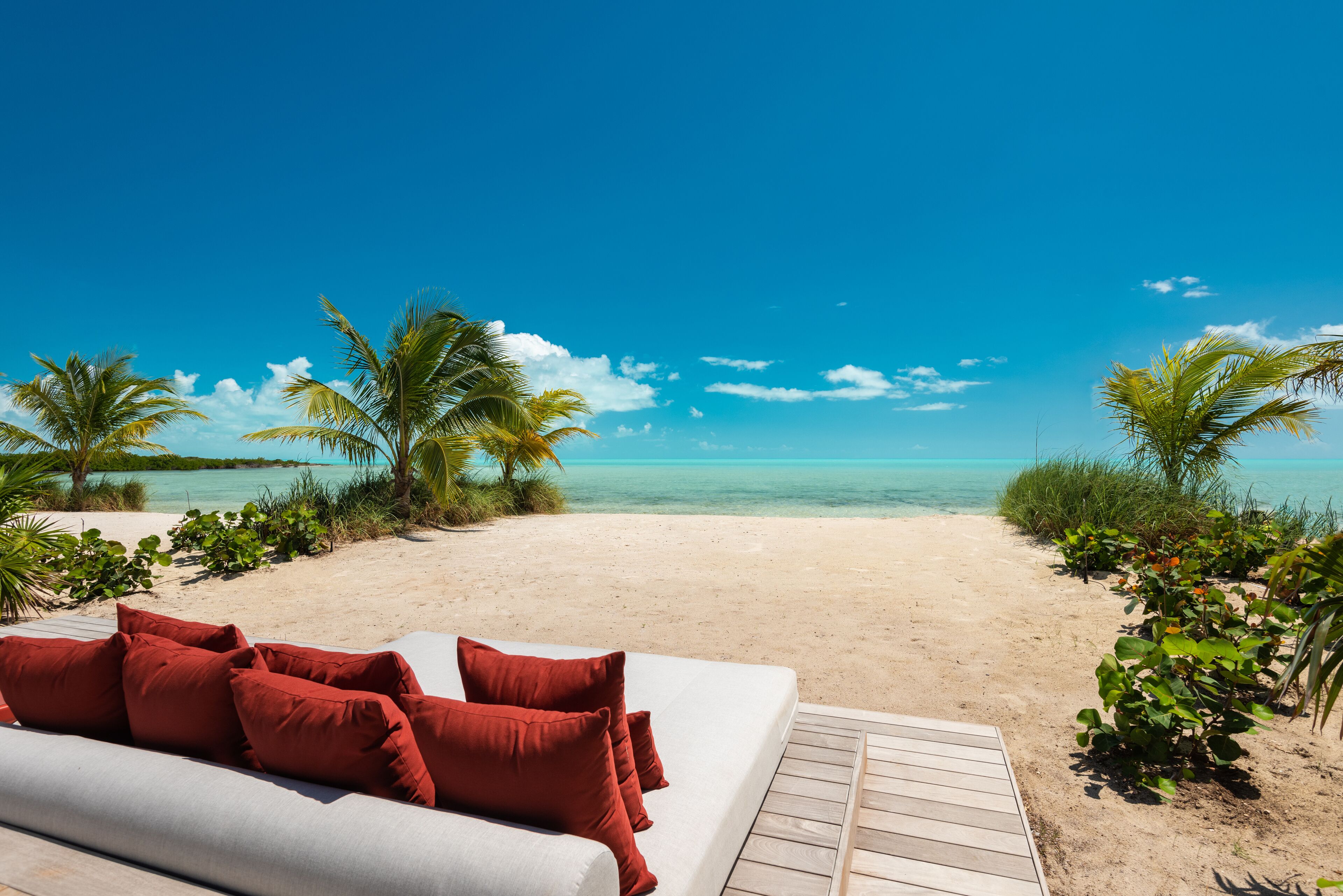 A beach view with a white couch and red pillows, palm trees, and a wooden deck.