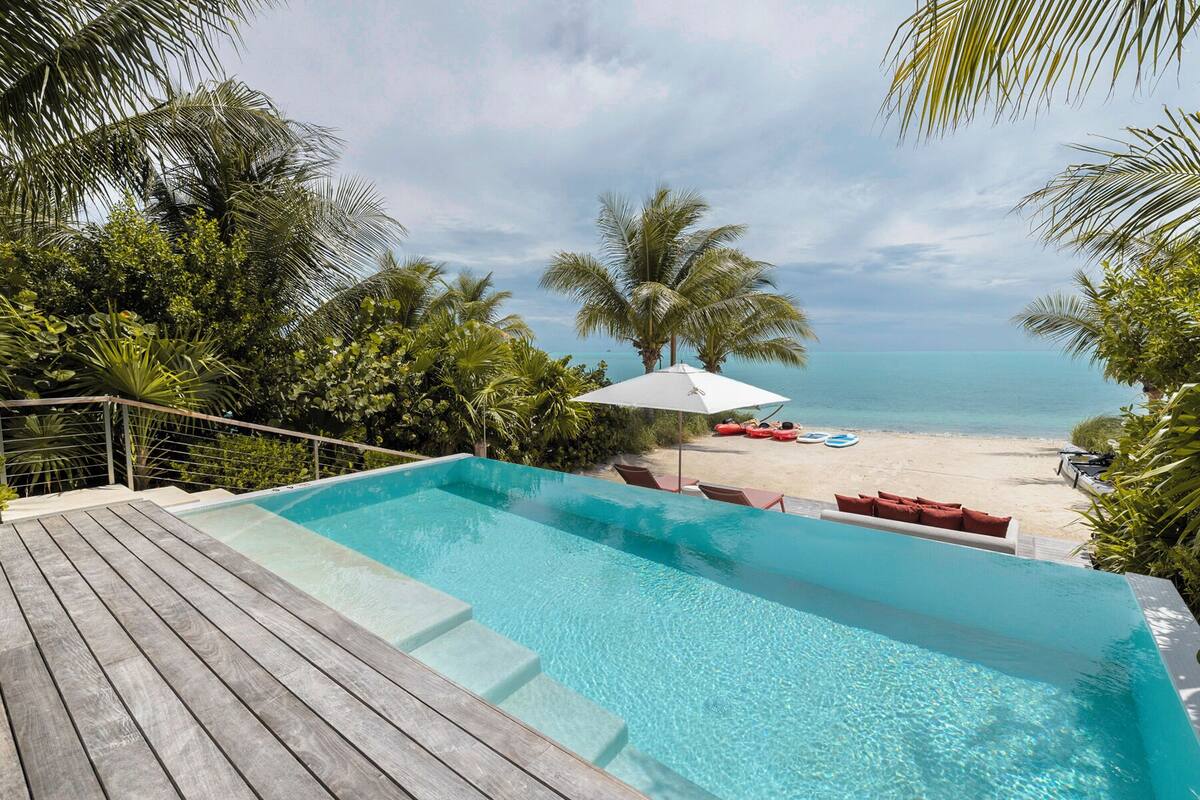A swimming pool with a wooden deck, a beach with lounge chairs and umbrellas, and a view of the ocean.