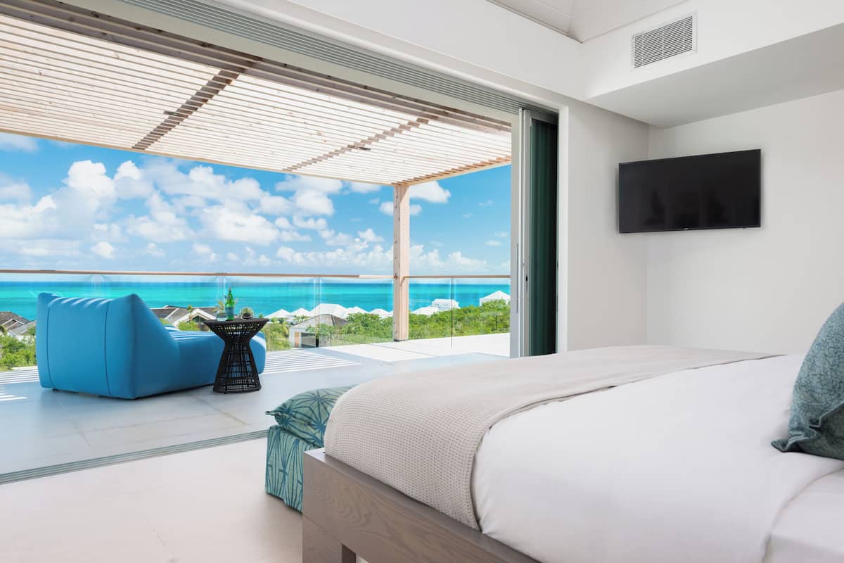 A bedroom with a large bed, a TV mounted on the wall, and a balcony with a view of the ocean.