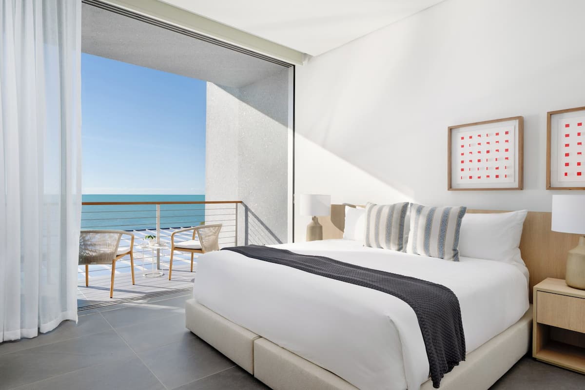 A modern hotel room with a large bed, a balcony with a view of the sea, and a minimalist decor.