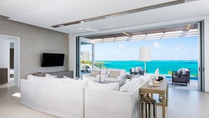 Un salon moderne doté d’un grand canapé d’angle blanc, d’un téléviseur à écran plat et d’un coin repas avec une table. La pièce est agrémentée de grandes portes vitrées donnant sur un balcon avec vue sur l’océan.