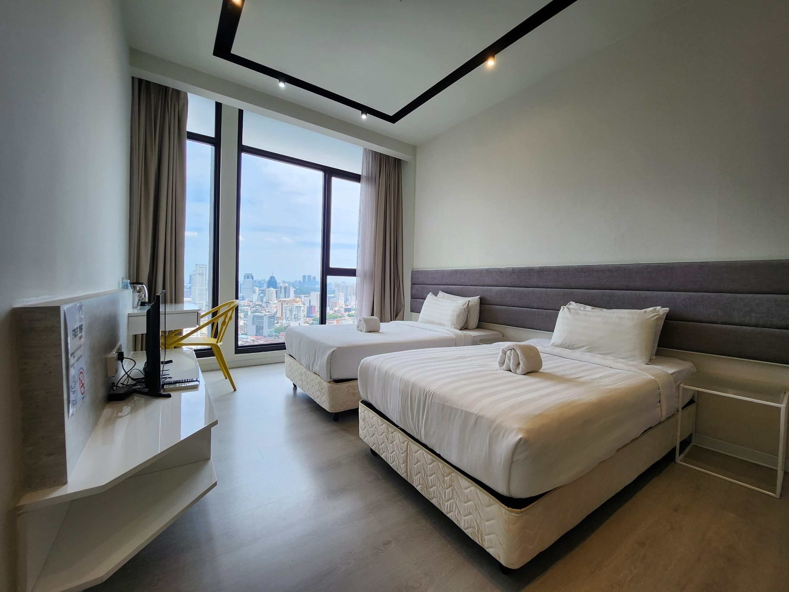 A modern hotel room with two beds, a desk with a computer, and large windows offering a city view.
