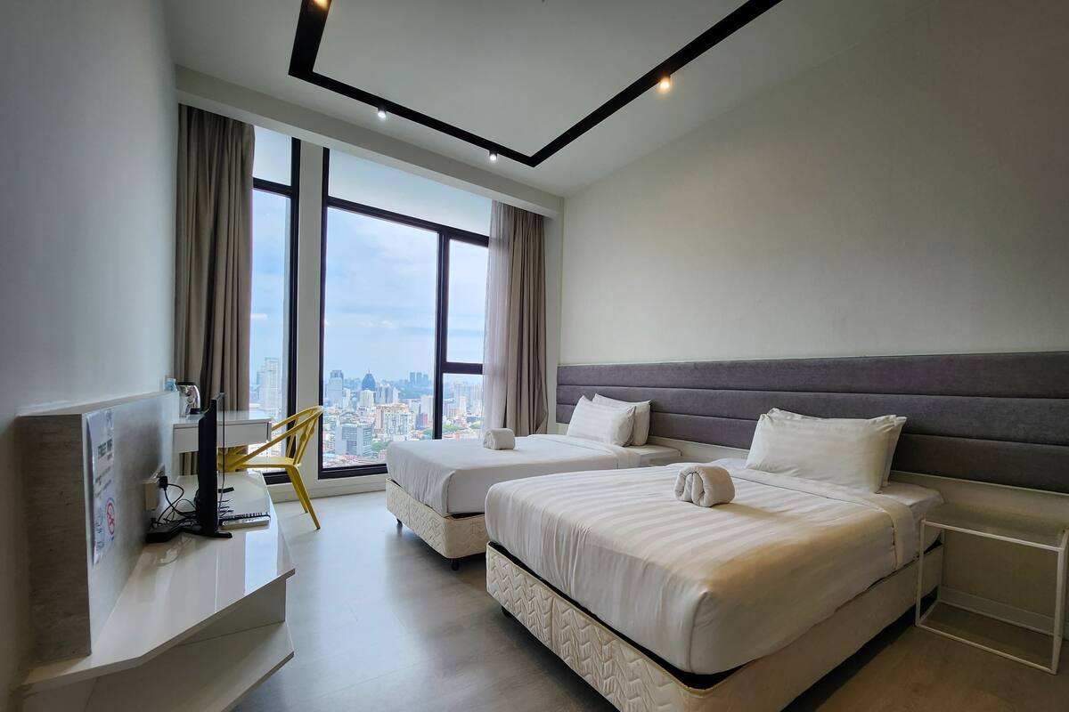 A modern hotel room with two beds, a desk with a computer, and large windows offering a city view.