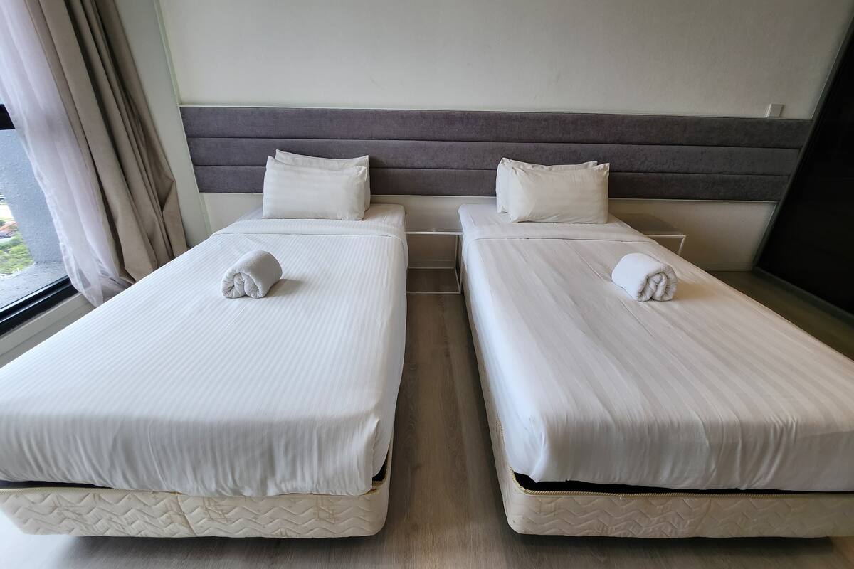 Two twin beds with white linens and pillows, a grey headboard, and a window with curtains.