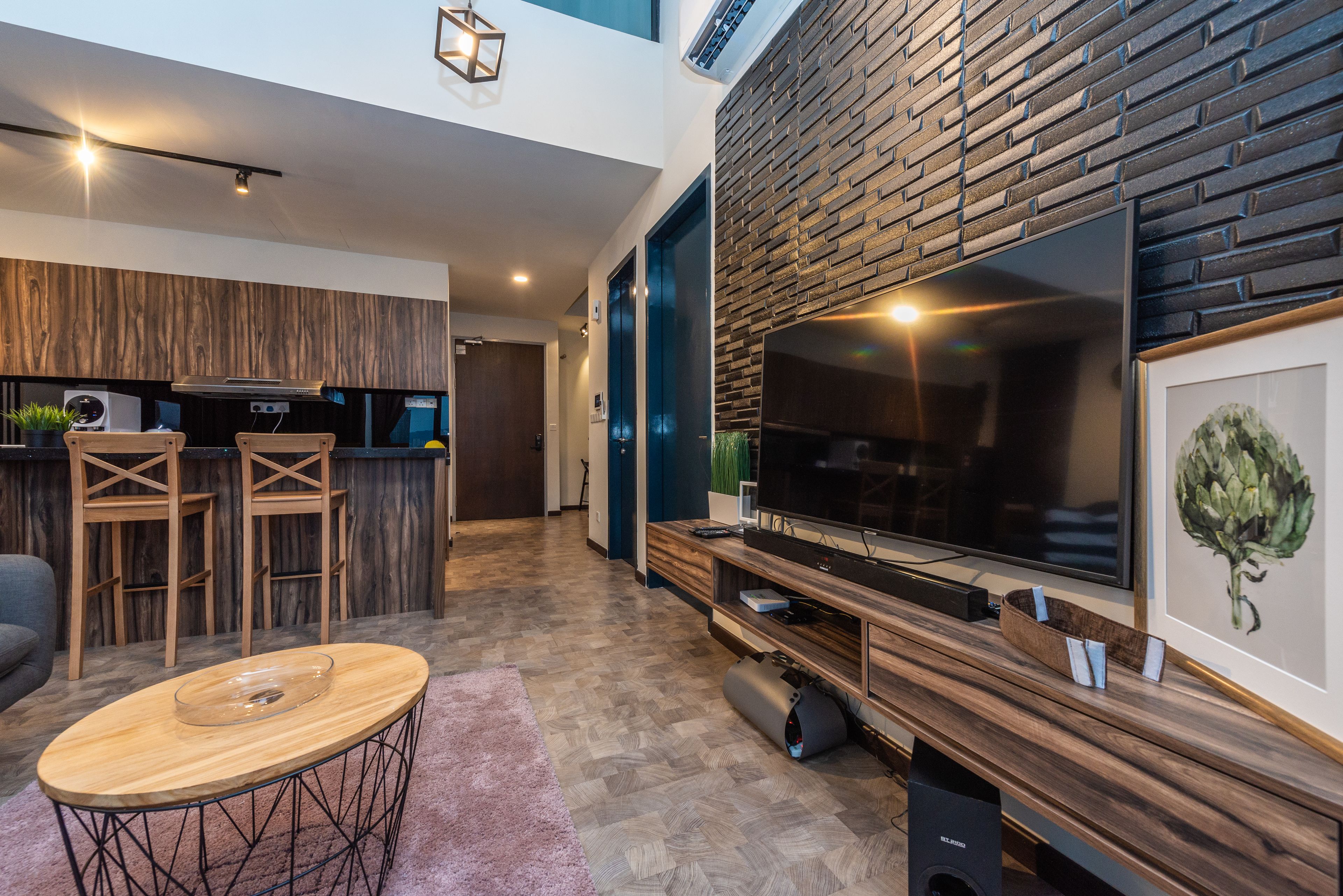 Ein modernes Wohnzimmer mit Holztisch, Fernseher auf einem Holzständer sowie einer Küchenzeile mit Holzwand und Barhocker.