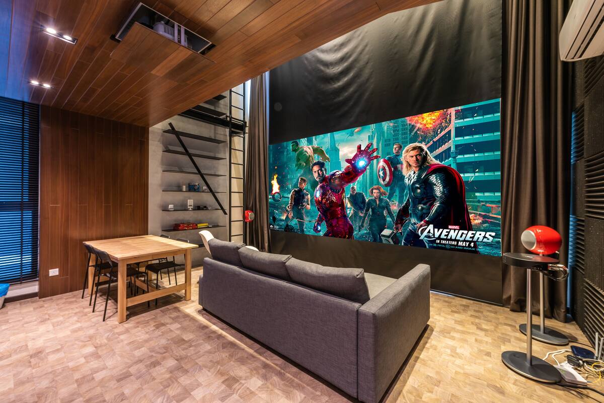 A modern living room with a large flat-screen TV displaying a movie poster, a wooden dining table, a grey sofa, and a red lamp.