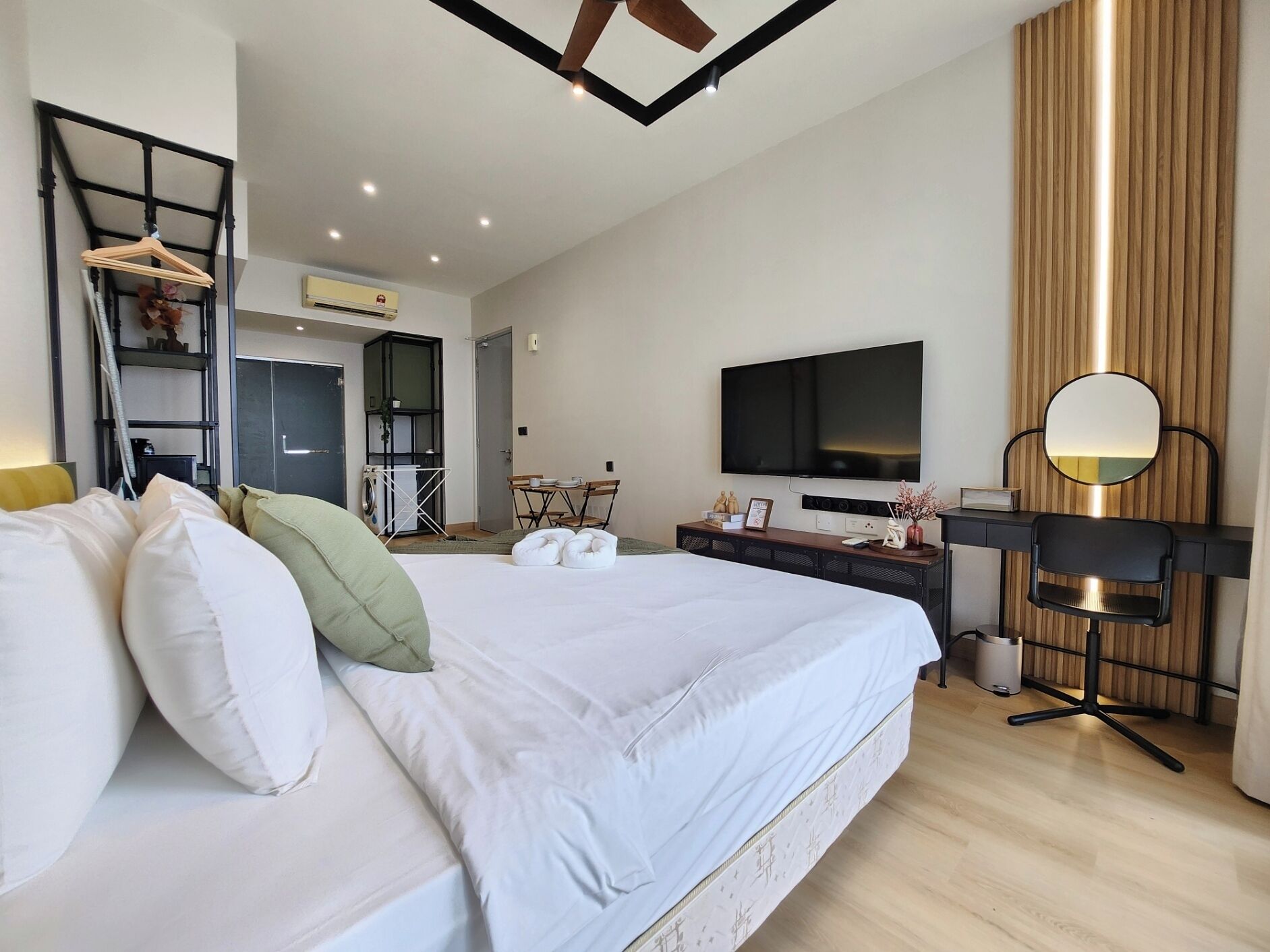 A modern hotel room with a large bed, a desk with a flat-screen TV, a mirror, and a kitchenette area.
