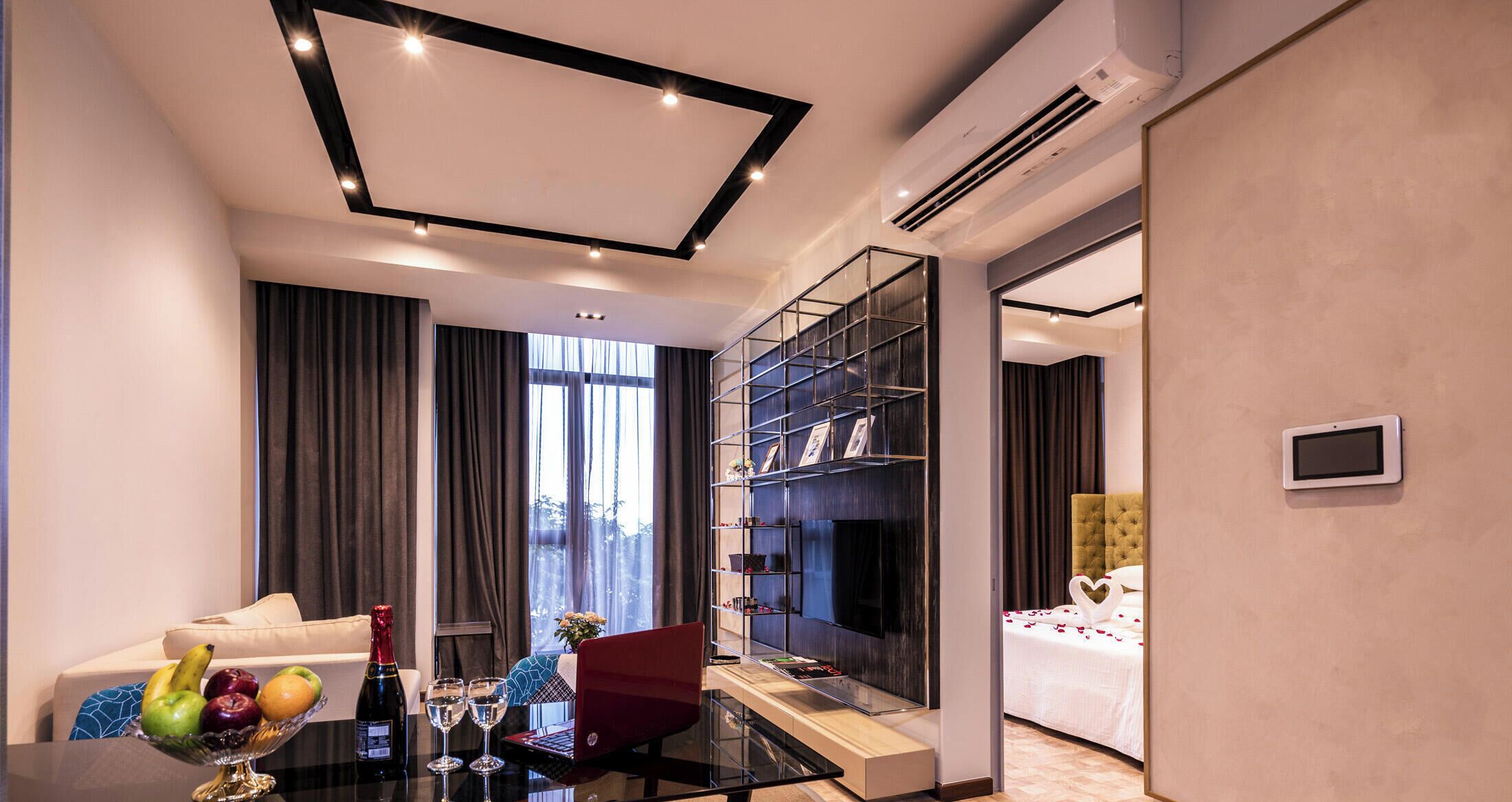 A modern hotel room with a glass dining table, a sofa, a red chair, a TV, and a bed with a white bedspread.