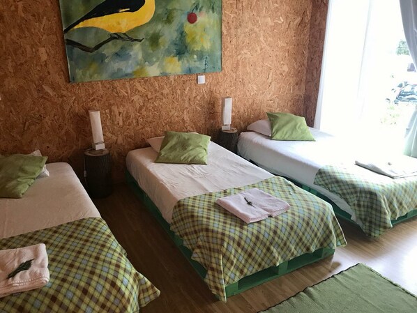 A room with three single beds, each with a green checkered bedspread and a white pillow. There is a painting on the wall above the beds, and a window with curtains to the right. - Biovilla Sustentabilidade (Palmela)