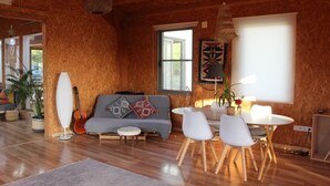 A cozy living room with a wooden interior, a sofa with patterned cushions, a dining table with chairs, and a hanging light fixture. - Biovilla Sustentabilidade (Palmela)
