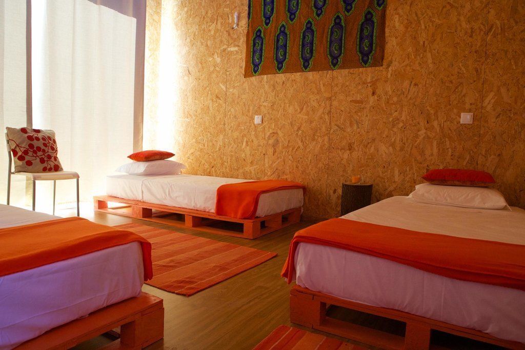 Ein Zimmer mit drei hölzernen Betten, jeweils mit weißer und orangefarbener Bettwäsche. Ein Stuhl mit rotem Kissen und ein dekoratives Wandbild über den Betten.