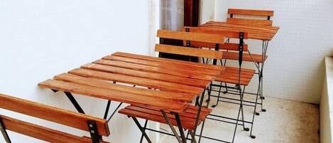 Una hilera de escritorios de madera con estructura metálica, ubicados en un pasillo estrecho con paredes blancas y una ventana con persianas.