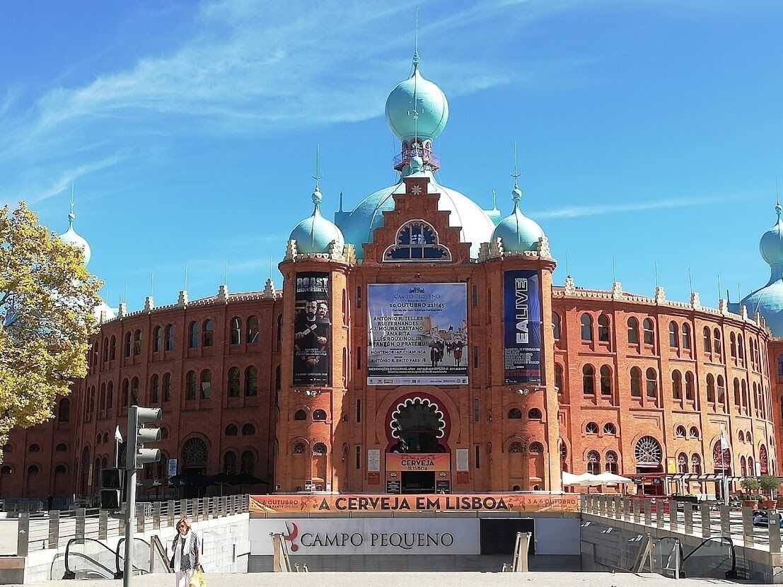 Un grand bâtiment en briques rouges, doté de plusieurs dômes et de fenêtres en arc de cercle, sur lequel on peut voir une enseigne bien visible indiquant « Cerveja Em Lisboa » et « Campo Pequeno ».