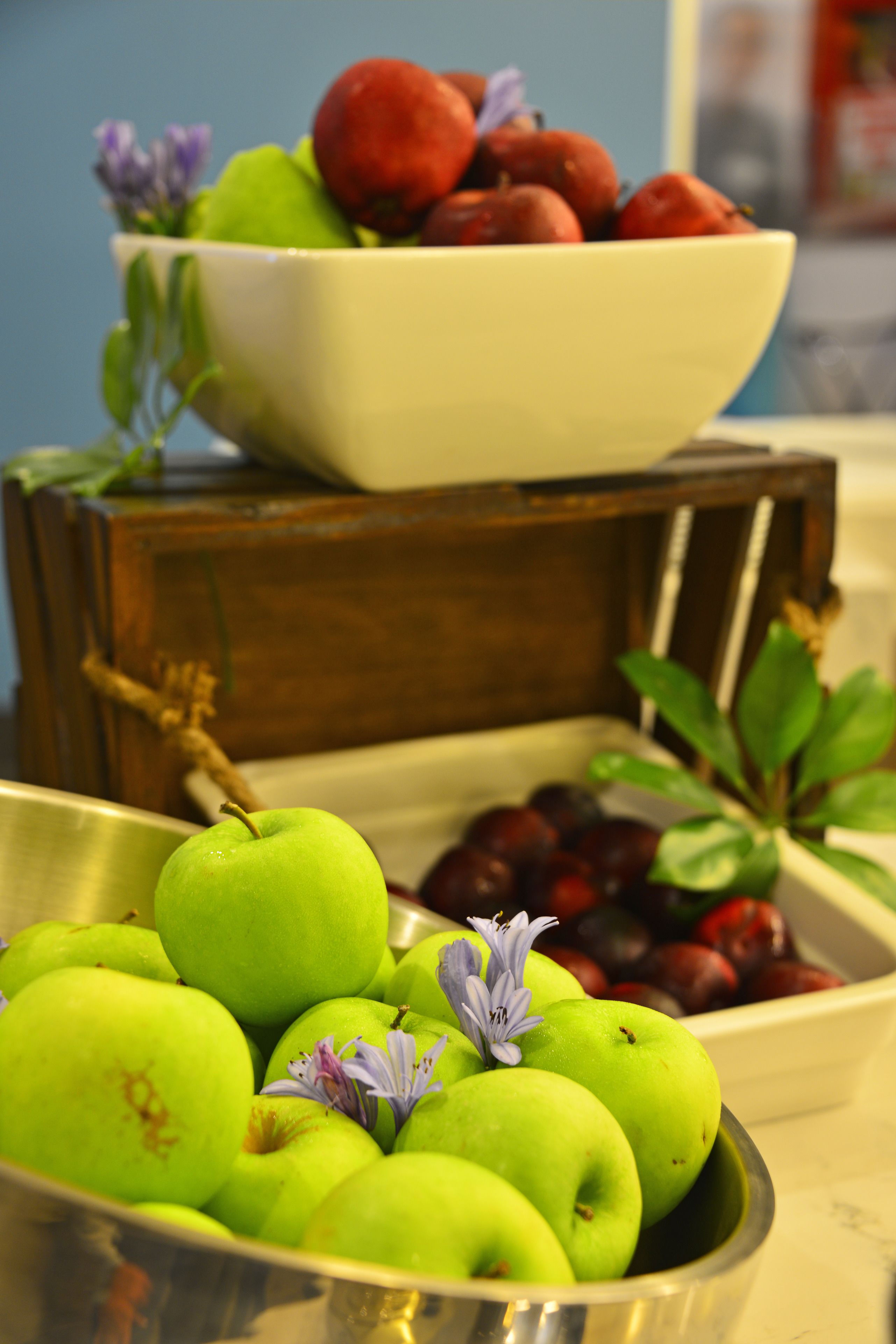 A bowl of green apples with a flower on top, a bowl of red fruits, and a wooden crate in the background.