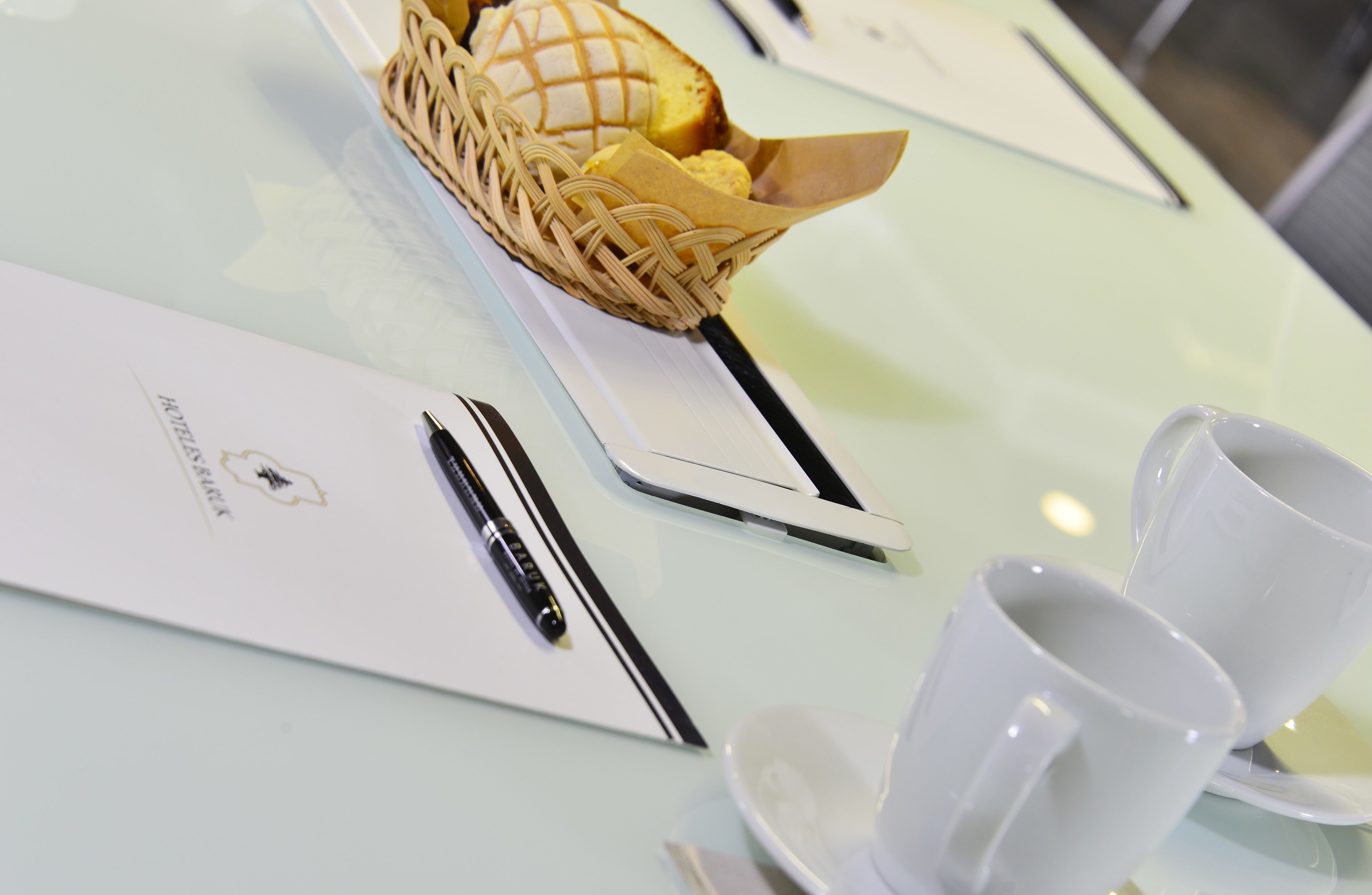 A table with a basket of bread, a pen, and two white cups on a glass table.
