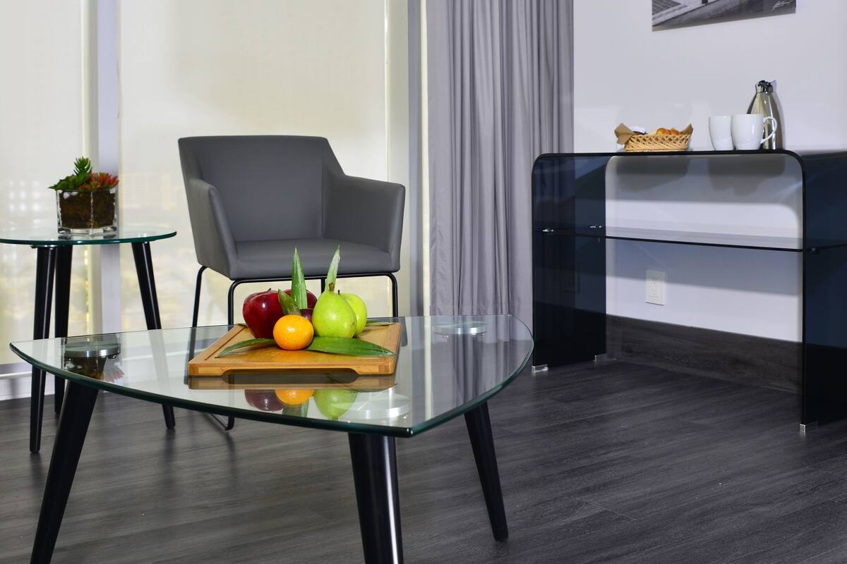 Ein modernes Hotelzimmer mit einem Glastisch, einem grau gepolsterten Sessel, einem schwarzen Beistelltisch, einem Glastischen und einem schwarzen Sofa.