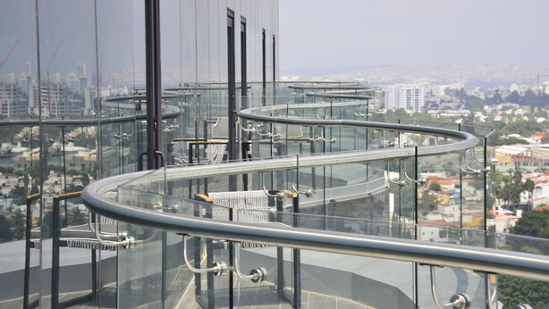 A modern glass balustrade with metal railings and wicker chairs on a high-rise balcony overlooking a cityscape.