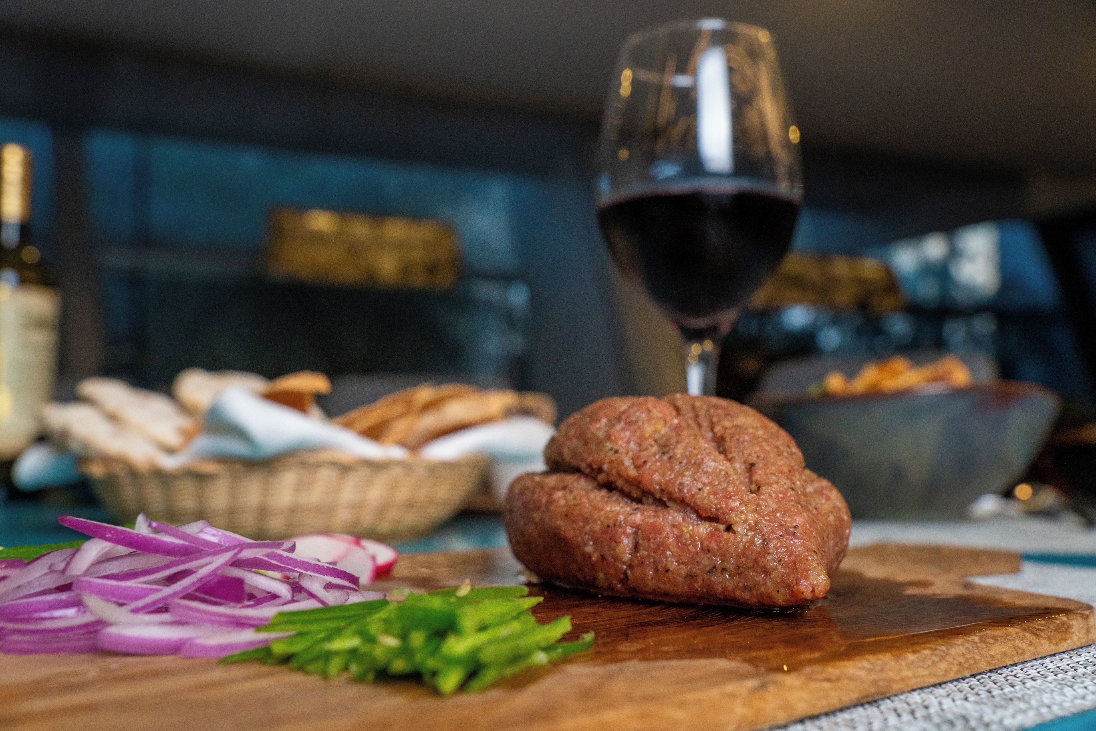 A wooden cutting board with a meat patty, sliced red onions, and green herbs.