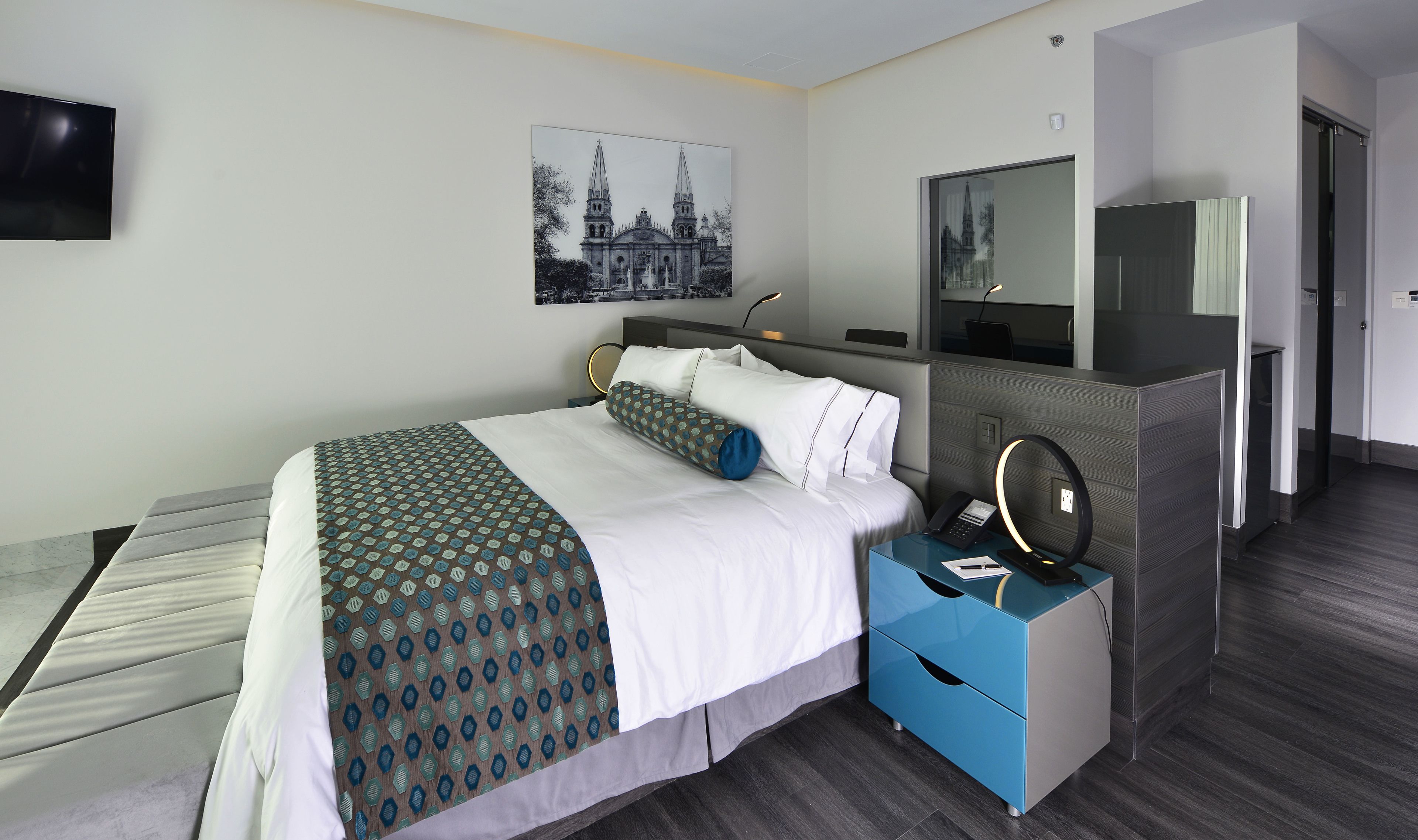 Une chambre d’hôtel avec un lit, une table de chevet avec un téléphone et une lampe, un miroir et un tableau représentant un bâtiment accroché au mur.