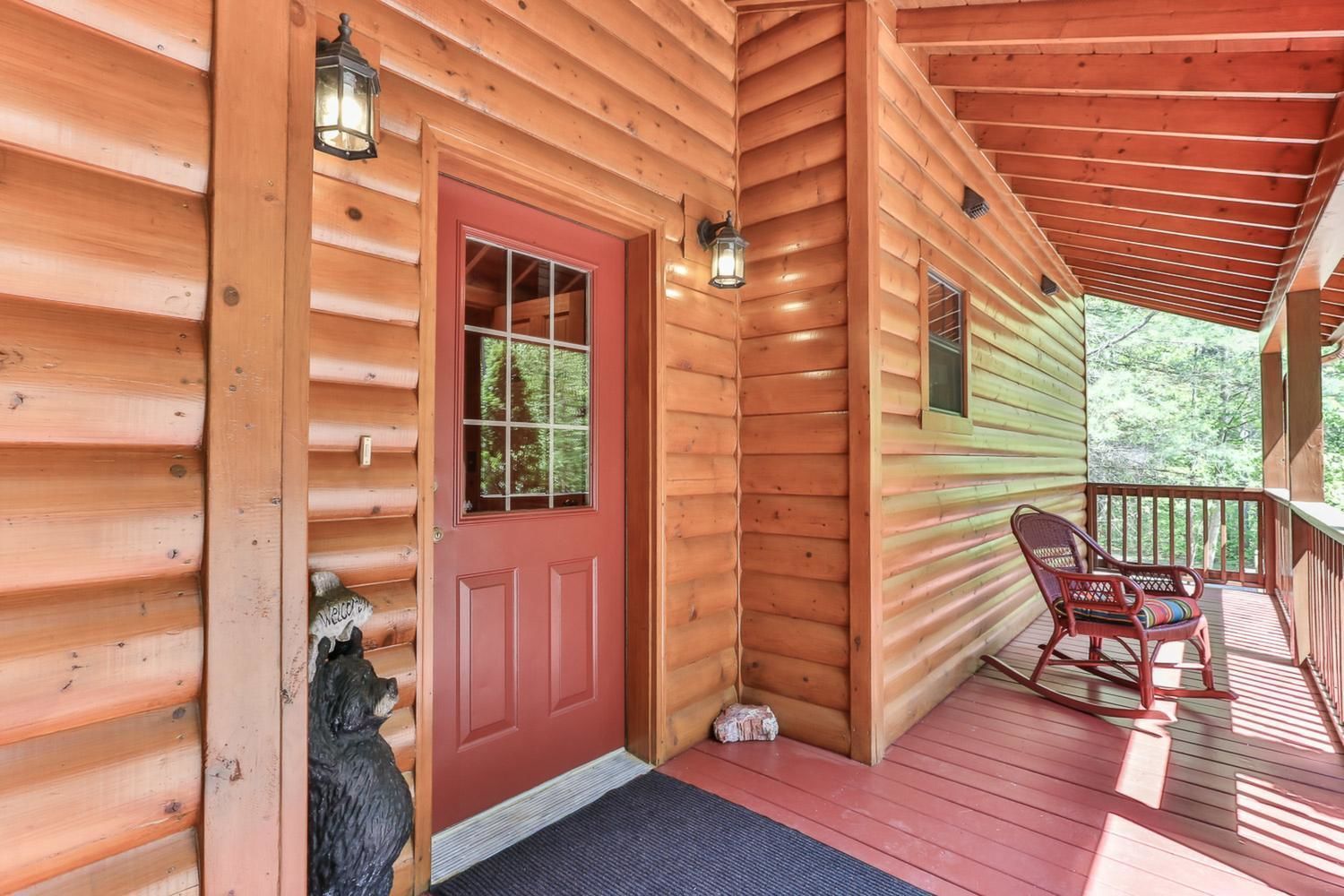 A wooden cabin with a red door, a porch with two rocking chairs, and a small dog statue outside.