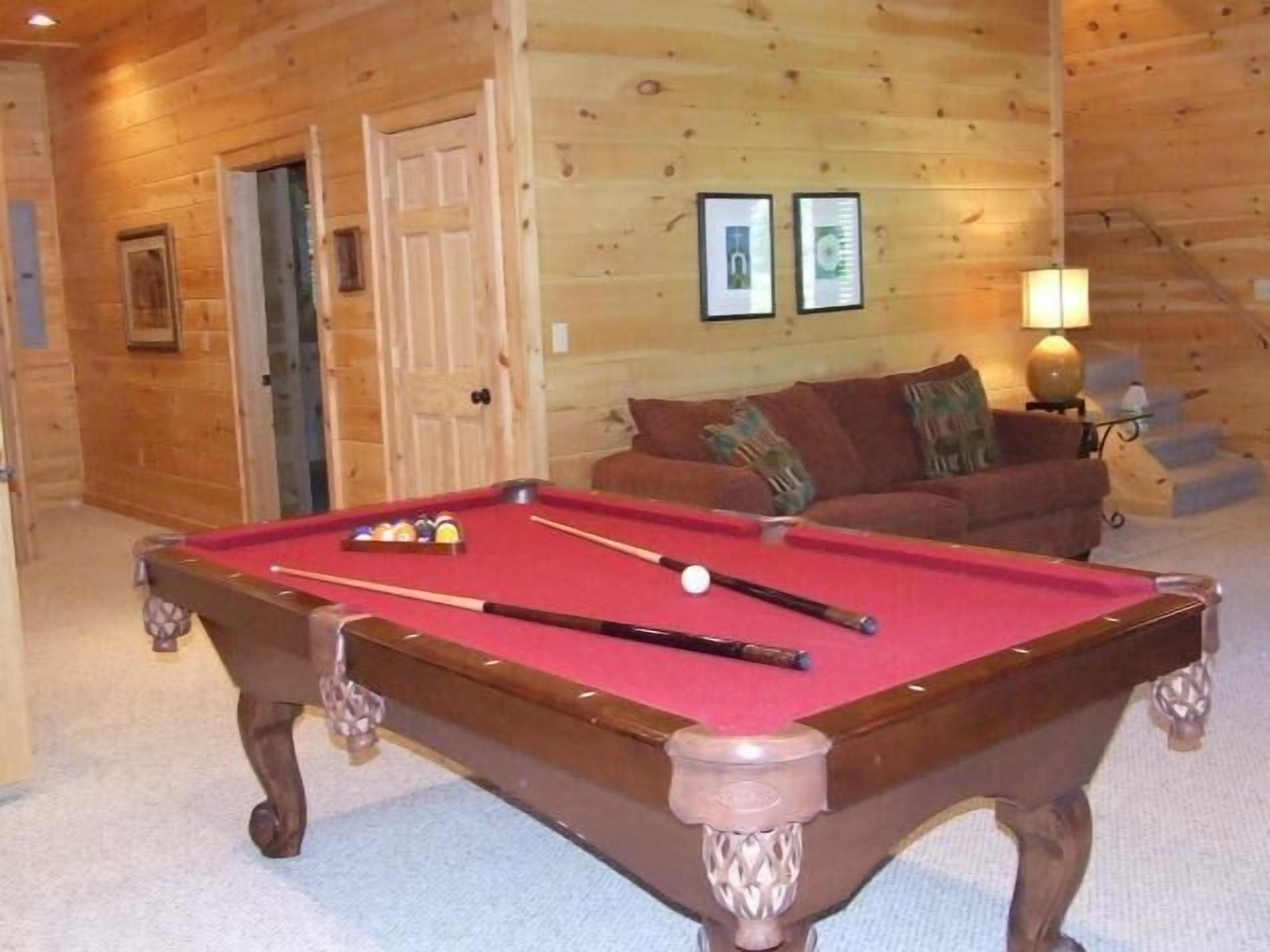 A pool table in a wooden room with a door, framed pictures, and a lamp.